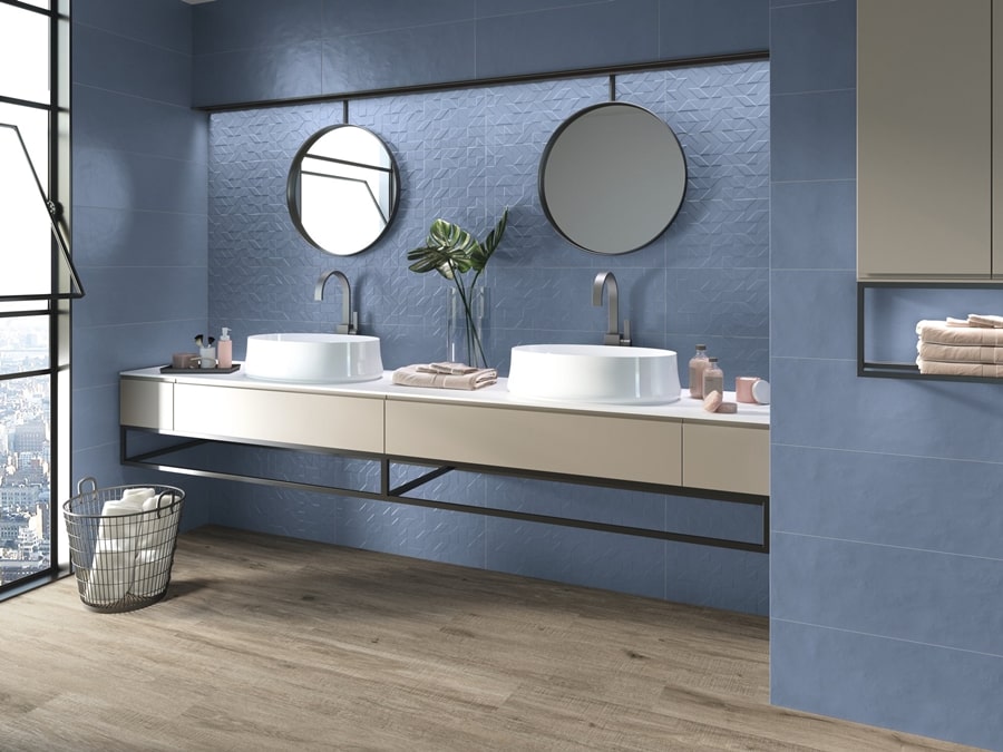 Łazienka z niebieskimi kaflami na ścianach i płytkami drewnopodobnymi na podłodze. Dwie białe umywalki.